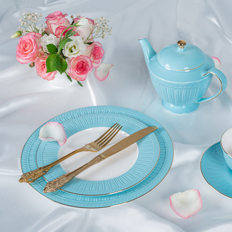 This image features a close-up of a blue bone china dinnerware set, elegantly paired with a bouquet of pink and white roses. The set includes a beautifully crafted teapot, a plate, and a cup, all adorned with a subtle blue glaze and intricate detailing.
