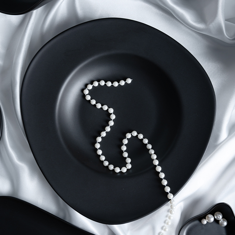 The image showcases a fine porcelain black plate, elegantly adorned with a string of lustrous pearls, set against a backdrop of luxurious white satin fabric.