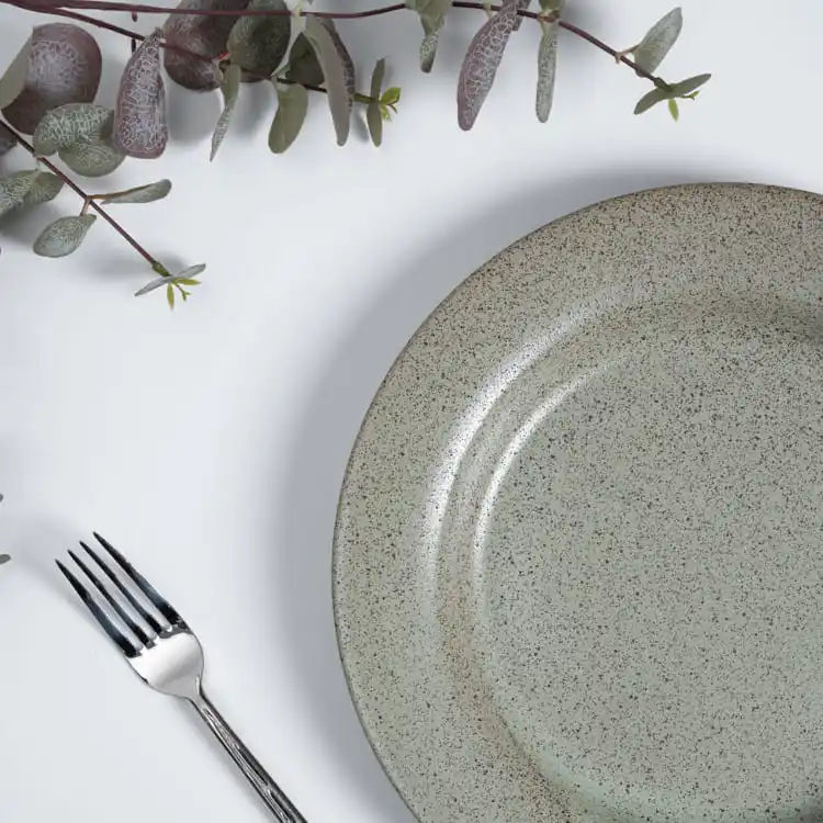 This image captures a single porcelain plate with a speckled glaze, set against a backdrop of eucalyptus leaves and a polished fork.