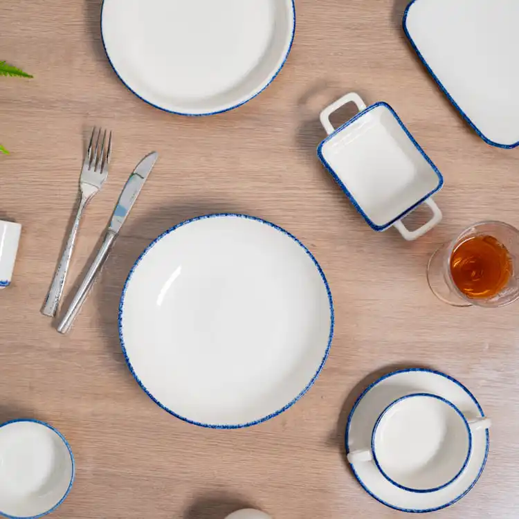 This image presents a beautifully arranged dining setup featuring modern white porcelain dinnerware. The set includes plates, bowls, and a small rectangular dish, all adorned with a delicate blue rim.