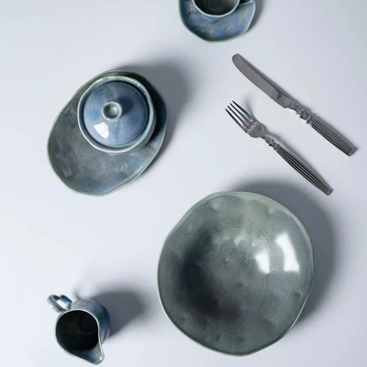 Esta imagen muestra una selección de vajilla de porcelana azul verdosa, que incluye un tazón, un plato pequeño, una taza y un platillo, complementados con un cuchillo y un tenedor.