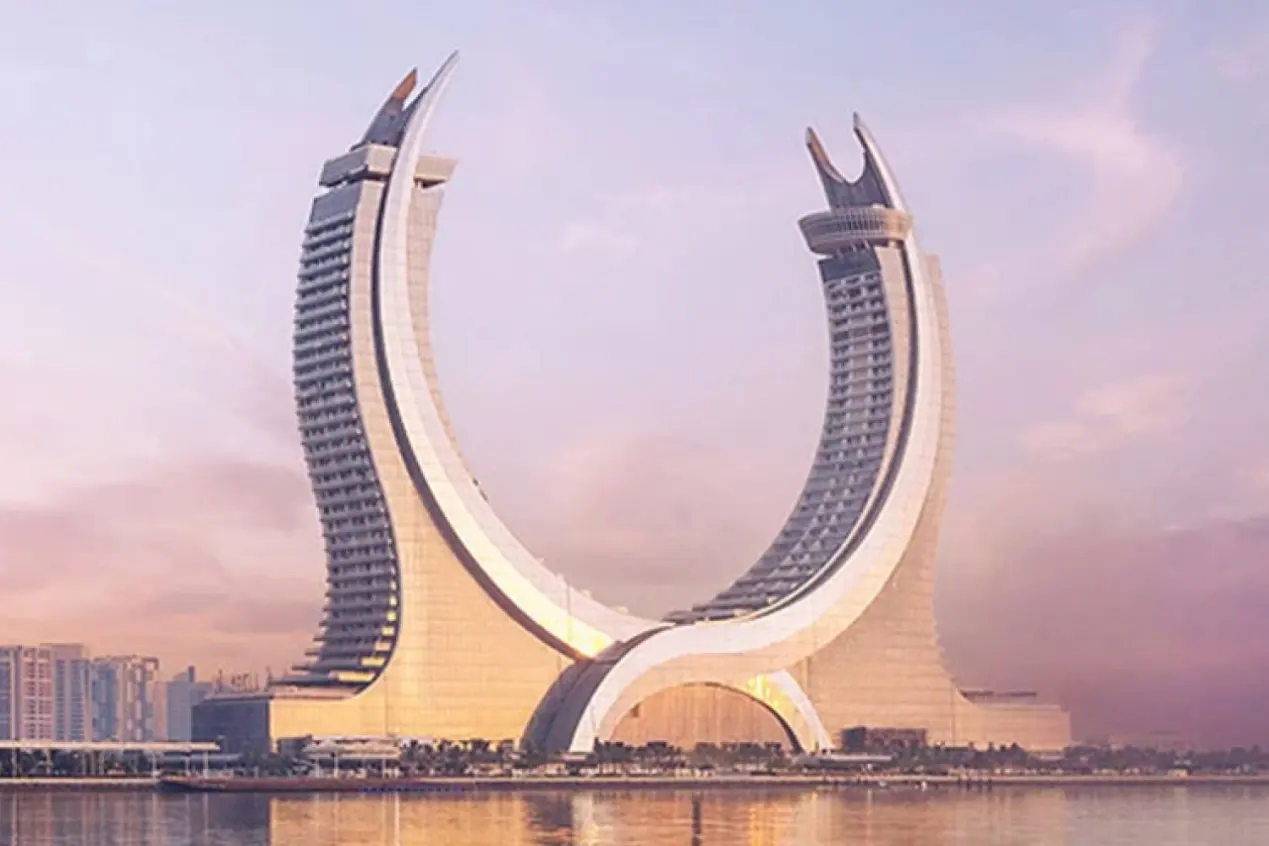Stunning waterfront exterior of Katara Towers Fairmont Hotel in Lusail, two curved crescent skyscrapers reflecting on calm water under soft pastel sunset sky.