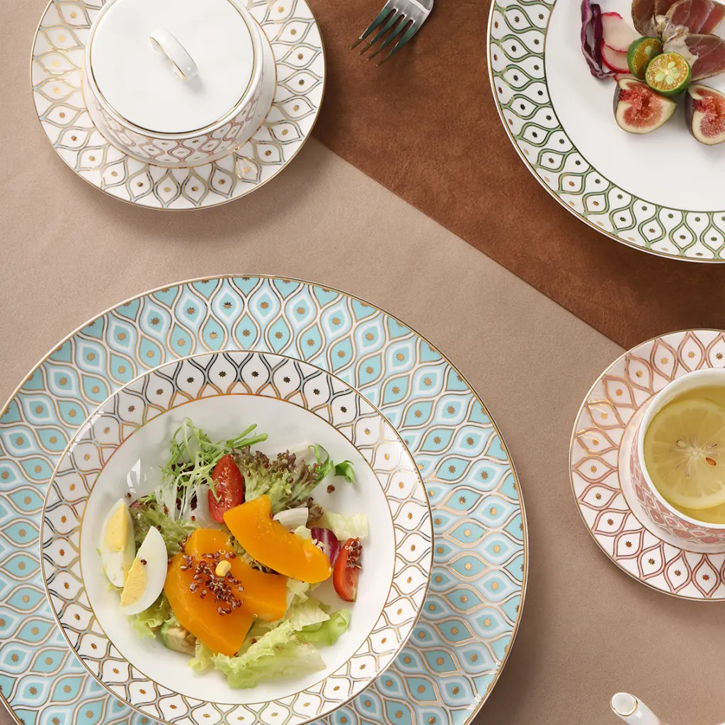 Top-down shot of patterned gold-rimmed bone china tableware laid with fresh salad, cured meat, lemon tea, teapot and covered soup bowl on neutral table mat.
