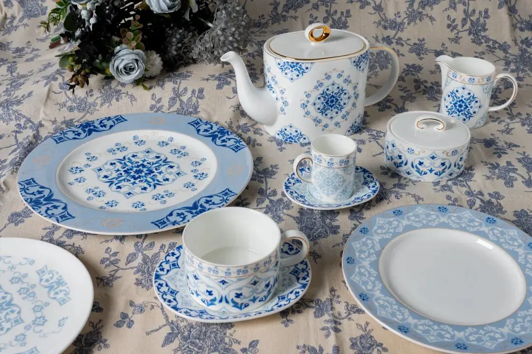 Blue and white porcelain dinnerware place setting with plates, teapot, cups on floral patterned tablecloth