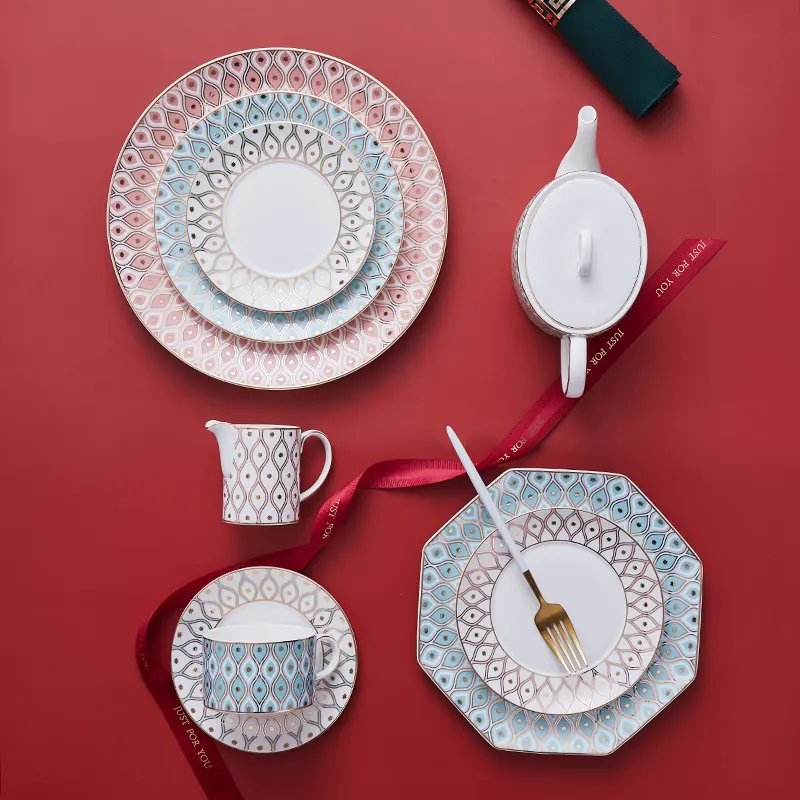 Flat lay of luxury bone china tea & dining set with gradient geometric patterns, teapot, plates, cup on vibrant red backdrop decorated with red satin ribbon.