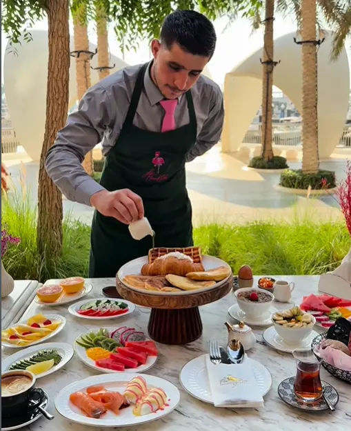 Waiter pouring syrup over waffles and pancakes served with fresh fruits on an outdoor dining table.