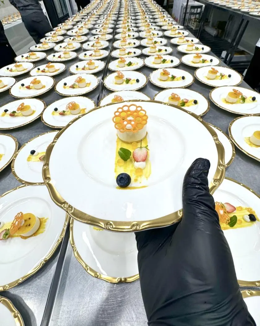 Gloved hand holding a custom fine china dessert plate with gold trim, showcasing plating for Versal's luxury banquet events.