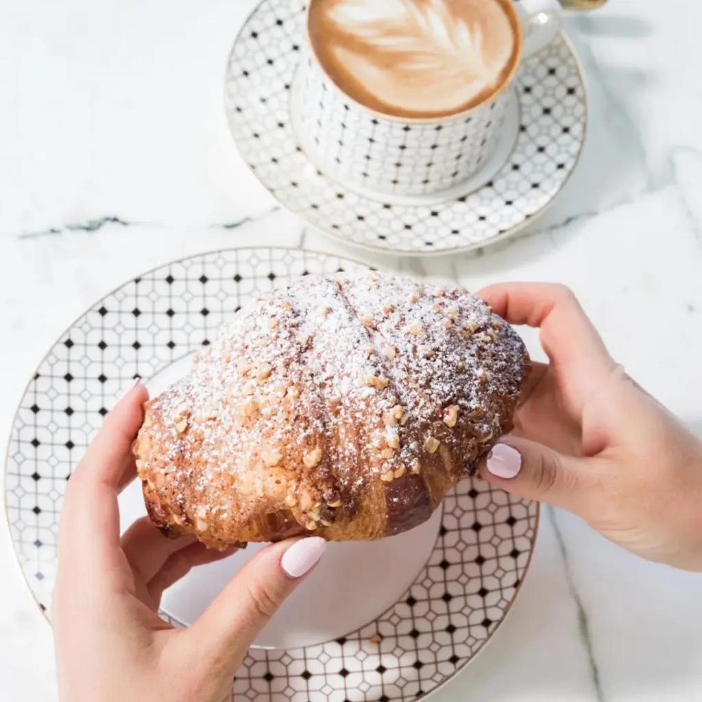 Hands holding a powdered croissant served on a PITO custom bone china dessert plate next to a cup of coffee.